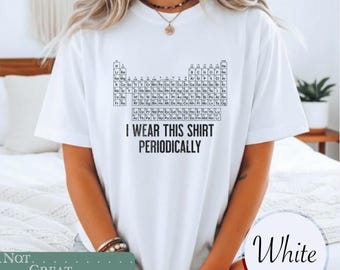 May include: A white short-sleeved t-shirt featuring a black graphic of the periodic table of elements printed on the chest. Below the table, the text reads I WEAR THIS SHIRT PERIODICALLY in bold black capital letters. The shirt has a crew neckline.