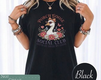 May include: A black t-shirt featuring a graphic of a white goose wearing sunglasses and a bow, surrounded by colorful flowers and butterflies. The text reads PORCH GOOSE SOCIAL CLUB and GOTTA GO DRESS MY GOOSE in a circular, decorative font.