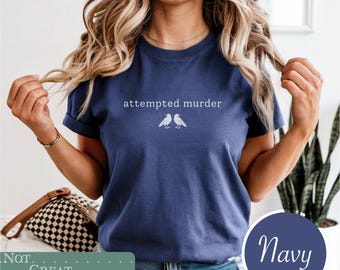 May include: A navy blue short-sleeve t-shirt featuring the white text "attempted murder" centered above a graphic illustration of two crows facing each other. The shirt has a crew neckline and a standard fit, showcasing a simple graphic design.