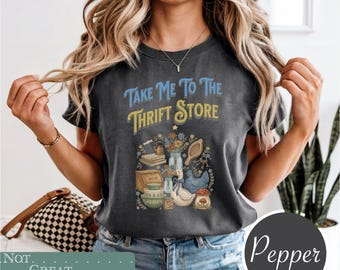 May include: A charcoal gray t-shirt featuring the text "Take Me To The Thrift Store" in blue and yellow. The graphic includes illustrations of a stack of books, a kettle, a bouquet of flowers, a wooden paddle, a bread box, bowls, and a goose.