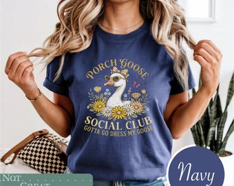 May include: A navy blue t-shirt featuring a graphic of a goose wearing a bow and sunglasses, surrounded by flowers and butterflies. The text reads: PORCH GOOSE SOCIAL CLUB, GOTTA GO DRESS MY GOOSE. The shirt has short sleeves and a crew neckline.