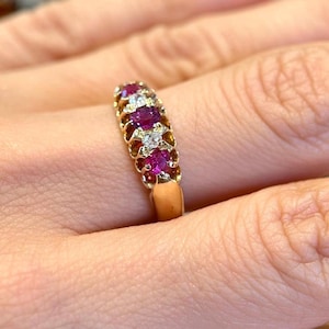 Puede incluir: Un anillo de oro con tres rubíes redondos de color rosa intenso que se alternan con dos pequeños diamantes redondos transparentes engastados en fila en la parte superior de la banda. El metal tiene un acabado pulido y estructura sólida.