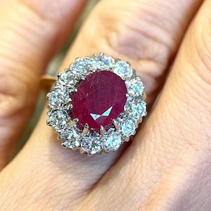 May include: A gold ring featuring a central oval-cut deep red ruby surrounded by a halo of twelve round brilliant-cut diamonds. The gemstone cluster is set on a polished gold band, creating a classic floral-inspired design with high clarity.