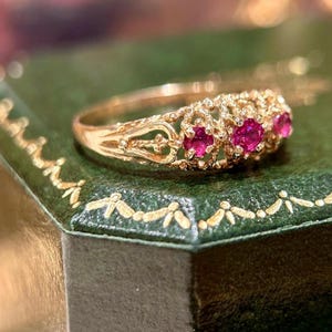 May include: A gold ring featuring three round, vibrant pinkish-red gemstones set in a delicate, openwork filigree band. The metalwork displays intricate, lace-like patterns surrounding each stone, creating a detailed and textured appearance.