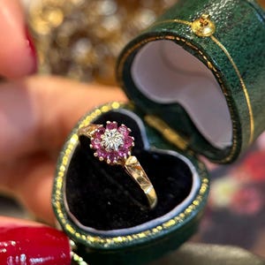 May include: A gold ring featuring a central diamond surrounded by a cluster of small pink rubies in a floral arrangement, presented inside a dark green heart-shaped jewellery box with a black velvet interior lining and gold trim detailing.