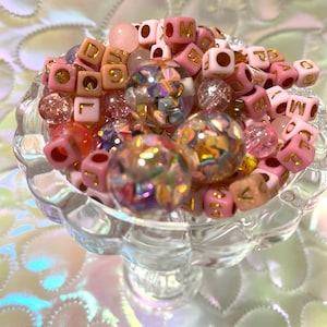 May include: A clear glass bowl filled with an assortment of beads. The beads include pink lettered cubes, iridescent spheres, and various other pink and clear beads. The bowl sits on a reflective surface.