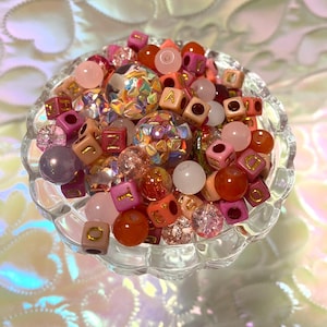 May include: A clear glass bowl filled with an assortment of colorful beads. The beads vary in size, shape, and color, including pink, orange, and clear beads. Some beads have gold lettered cubes. The bowl sits on a reflective surface.