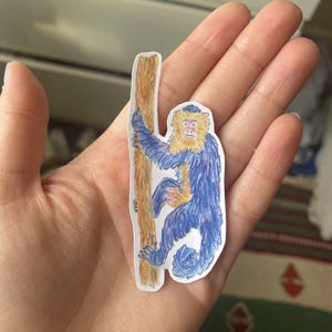 May include: A die-cut sticker featuring a hand-drawn illustration of a monkey with blue fur and a yellow mane climbing a brown tree trunk. The monkey has a light-colored face and is depicted in a vertical orientation with a curled tail at the bottom.