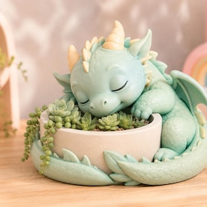 Può includere: Una statuetta di drago verde menta chiaro avvolta attorno a un vaso cilindrico. Il drago ha gli occhi chiusi, piccole corna e squame strutturate. Il vaso contiene piccole piante grasse, tra cui una pianta a collana di perle.