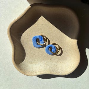 May include: A pair of blue and white marbled earrings with gold-colored hoops. The earrings are square-shaped with a rounded inner circle. They are displayed on a beige, organic-shaped tray.