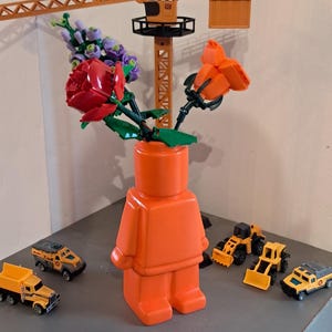 Könnte beinhalten: Eine orangefarbene Lego-Figur-Vase hält rote und lila Lego-Blumen. Miniatur-gelbe Baufahrzeuge und ein Kran sind um die Vase auf einer grauen Oberfläche angeordnet. Die Szene ist verspielt und farbenfroh.