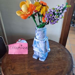 Peut inclure: Un vase en céramique bleu et blanc en forme de figurine contient des fleurs en plastique colorées. Les fleurs sont orange, jaunes et violettes. Une carte rose avec le mot "Céramique" est sur la table.