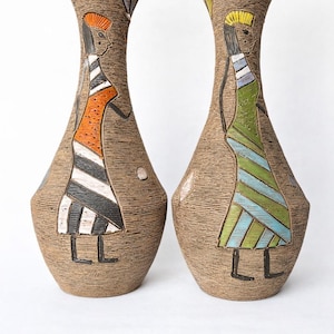 Può includere: Due vasi decorativi in ceramica con superfici marroni strutturate. Ogni vaso presenta una figura dipinta con un abito colorato. Una figura indossa arancione e nero, l'altra verde e blu. I vasi hanno un aspetto unico, fatto a mano.