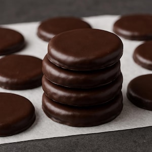 May include: A stack of six round chocolate-covered cookies, with several more scattered around on a white surface. The cookies have a shiny, dark brown coating and a smooth texture. The background is a dark gray.