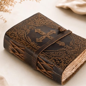 Puede incluir: Un diario de cuero marrón oscuro con una cruz en relieve y complejos motivos florales en la portada. El libro se asegura con una correa de cuero y presenta un cordón lateral de cuero, junto con páginas de papel grueso y texturizado.