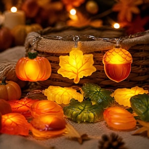 May include: A string of LED fairy lights featuring translucent, glowing autumn-themed shapes including orange pumpkins, yellow maple leaves, and brown acorns. The lights are arranged in a decorative cluster, emitting a warm, soft light.