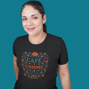 May include: Black t-shirt with a floral design and the text "CAFÉ Y CHISME" in orange and teal. The design is surrounded by orange, teal, and white flowers. The shirt is a crew neck.