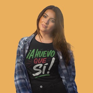 Può includere: Una maglietta nera con la scritta "¡A HUEVO QUE SÍ!" in verde, rosso e bianco. La persona indossa una camicia a quadri blu e bianca sopra la maglietta e pantaloni neri. Lo sfondo è di colore arancione.