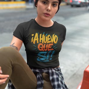 Può includere: Top corto nero con la scritta "¡A HUEVO QUE SI!" in giallo, arancione e blu. La maglietta ha un girocollo e maniche corte. La persona indossa pantaloni verde oliva e una camicia a quadri legata in vita.