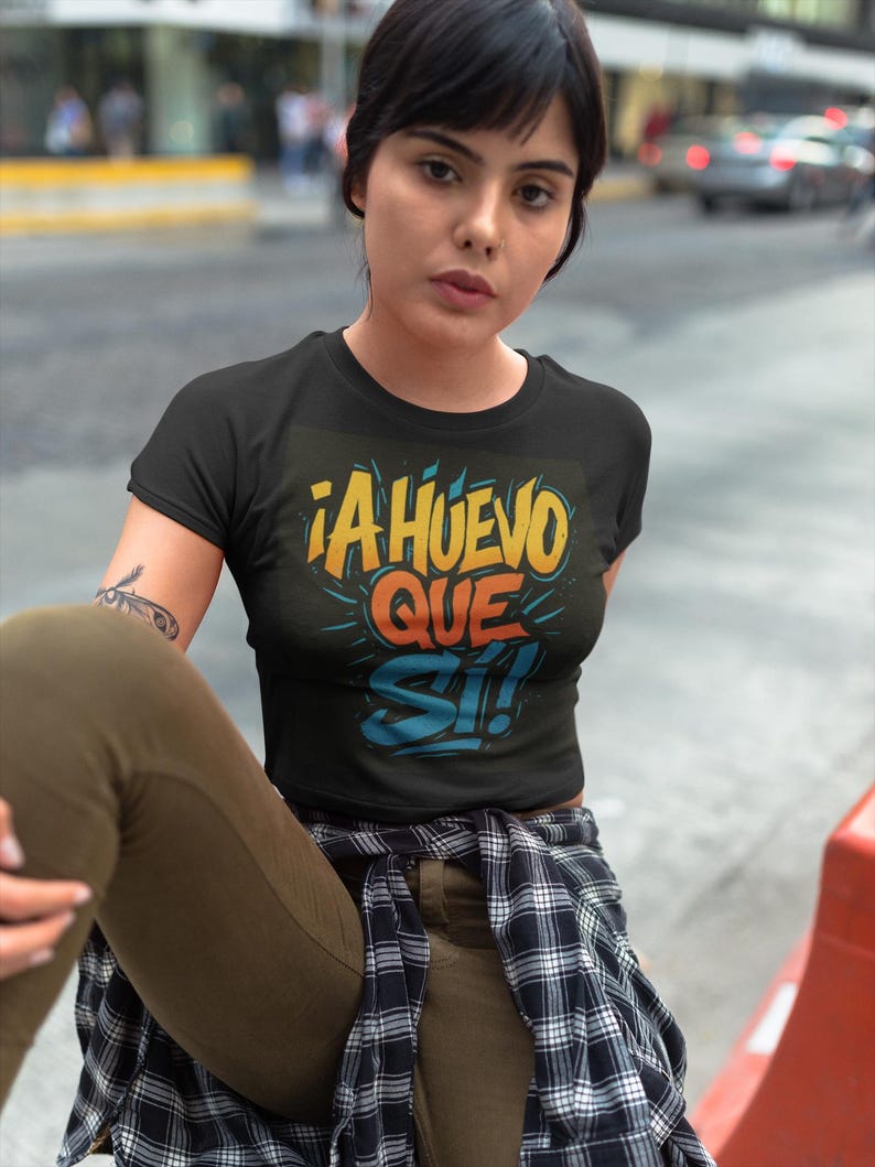 Puede incluir: Top corto negro con el texto "&iexcl;A HUEVO QUE SI!" en amarillo, naranja y azul. La camiseta tiene cuello redondo y mangas cortas. La persona lleva pantalones verde oliva y una camisa a cuadros atada a la cintura.