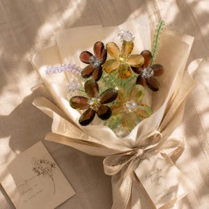 May include: A bouquet of faceted crystal flowers in shades of amber, brown, and clear, accented with green beaded stems and lavender sprigs. The flowers are wrapped in beige paper with a ribbon and a tag that reads Especially for you WISH YOU ALL THE BEST.