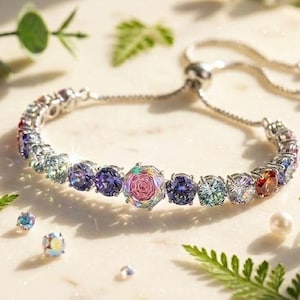 May include: A silver bracelet with a row of colorful gemstones. The bracelet features a central rose-shaped gem and is surrounded by various colored stones, including purple, blue, and red. The bracelet is displayed on a light-colored surface with greenery.