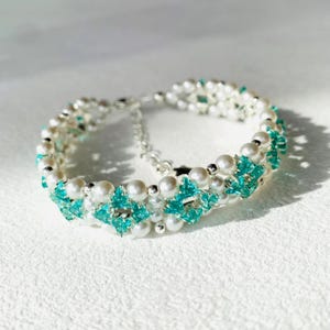 Puede incluir: Una pulsera delicada con perlas blancas y cristales turquesa brillantes. La pulsera está adornada con pequeñas cuentas plateadas y un cierre de cadena plateada. El diseño es simétrico, con los cristales dispuestos en un patrón repetitivo.