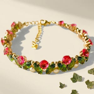 Puede incluir: Un brazalete dorado con piedras preciosas rojas y verdes, pequeñas perlas blancas y un dije en forma de corazón. El brazalete presenta un diseño delicado con motivos florales y de hojas, con una longitud aproximada de 18 cm.