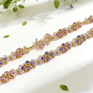 Puede incluir: Dos collares dorados con diseños florales. Uno presenta piedras preciosas rosas y moradas, el otro un diseño de rama con gemas rosas. Los collares están sobre un fondo blanco, con hojas verdes.