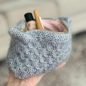May include: A hand-knitted, light gray cosmetic bag with a gold zipper. The bag is filled with makeup products. The interior lining is a pink and white striped fabric. The bag is made of a soft, textured yarn.