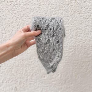 May include: A gray knitted sleeve held against a white textured wall. The sleeve has a diamond-shaped openwork pattern. The knitted item appears soft and is likely made of wool or a similar material. The sleeve is a light gray color.