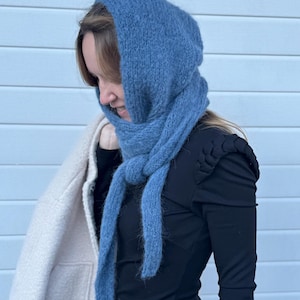 May include: A blue knitted headscarf with a fuzzy texture, featuring a hood-like design that covers the head and ties securely under the chin with two long, dangling ends. The knit fabric has a soft, slightly brushed appearance and a solid blue color.