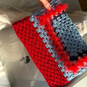 Puede incluir: Una funda de portátil de ganchillo hecha a mano en rojo y azul. La funda está dividida diagonalmente, con rojo en la parte inferior izquierda y azul en la parte superior derecha. Un ribete rojo y esponjoso acentúa la costura diagonal. La funda está diseñada para un portátil.