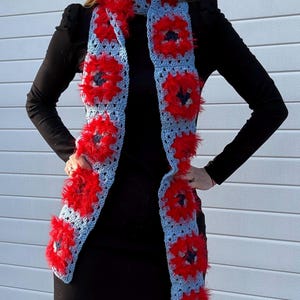 May include: A handmade crochet scarf featuring a light blue base with red floral accents. The scarf has a rectangular shape with a repeating pattern of red flowers and a fluffy texture, creating a unique and eye-catching accessory.