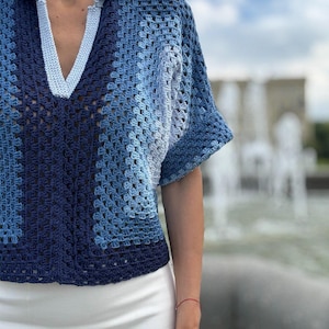 Op de afbeelding: Een gehaakte top met korte mouwen in een verloop van blauwtinten, variërend van diep marineblauw tot licht hemelsblauw. Het kledingstuk heeft een V-hals met een effen lichtblauwe geribbelde kraag en een vierkant patroon over het hele lijfje.
