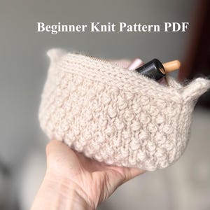 May include: A beige, hand-knitted pouch with a textured, bobble stitch design. The pouch is open, revealing a glimpse of cosmetics inside. The text "Beginner Knit Pattern PDF" is displayed above the pouch.