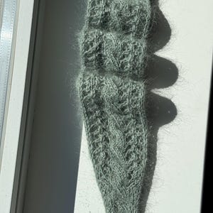 Könnte beinhalten: Ein salbeigrünes Strickstirnband mit einem strukturierten Zopfmuster. Das Accessoire ist aus einem flauschigen, weich wirkenden Garn gefertigt und weist ein markantes, vertikal verlaufendes Flechtmuster auf, das für eine warme und haptische Optik sorgt.