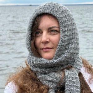 May include: A grey knitted balaclava hood with a textured ribbed pattern. The headpiece features a fitted hood that frames the face and extends into two long, scarf-like ends that drape downwards, crafted from a soft, fuzzy yarn material.