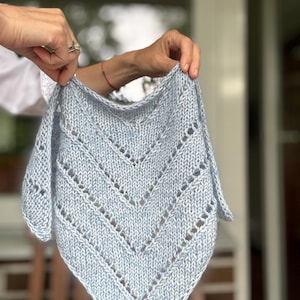 May include: A light blue triangular knit shawl featuring a series of chevron-shaped eyelet lace patterns. The garment is crafted from a soft, heathered yarn with a visible stockinette stitch texture, creating a pointed bottom edge and a wide top.