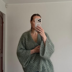 May include: A sage green, open-front cardigan with a loose, open-weave design. The cardigan features wide, bell sleeves and a V-neckline. The soft, textured knitwear is a cozy, stylish garment.