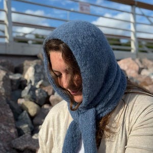 Könnte beinhalten: Ein staubblauer, gestrickter Kapuzenschal mit einer texturierten, flauschigen Oberfläche. Das Accessoire besteht aus einer abgerundeten Kopfbedeckung, die in zwei lange, herabhängende Enden übergeht, gefertigt aus weichem, dickem Garn.