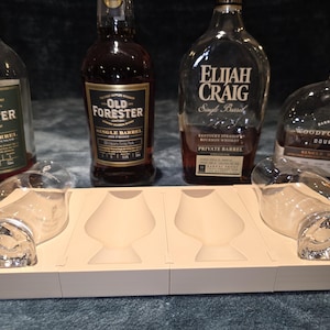 May include: Four bottles of bourbon whiskey, including Old Forester and Elijah Craig, are displayed with two whiskey glasses and a beige tray. The bottles have various labels and are partially filled.