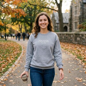 May include: A gray sweatshirt with the text "DESIGN YOUR OWN [CUSTOM TEXT HERE] FALL 2024" in white. The sweatshirt is paired with blue jeans. The person is holding a reusable coffee cup and walking on a path with fall foliage.