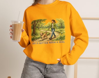 May include: A bright orange crewneck sweatshirt featuring a graphic illustration of a person walking a golden dog on a path through a park. The text below the illustration reads, "LIFE IS BETTER WITH A DOG" in a dark, capitalised sans-serif font.