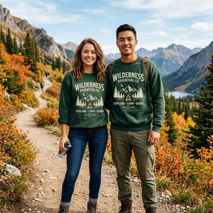 May include: Two people wearing matching dark green sweatshirts with the text "Wilderness Adventure Co." and a mountain and campfire graphic. The sweatshirts are paired with jeans and cargo pants. The background features a mountain landscape with a hiking trail.