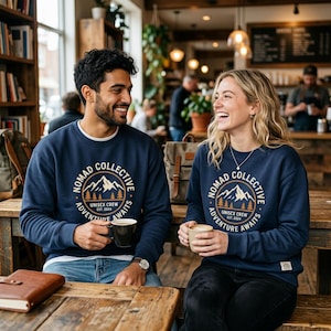May include: Two people are seated at a wooden table, each wearing a navy blue sweatshirt. The sweatshirts feature a circular graphic with a mountain, trees, and the text "NOMAD COLLECTIVE, UNISEX CREW EST. 2024, ADVENTURE AWAITS". They are holding mugs.