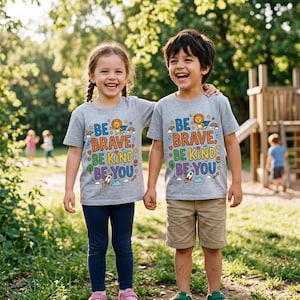 Può includere: Due bambini indossano magliette grigie con le parole "BE BRAVE, BE KIND, BE YOU" in lettere colorate. Le magliette presentano un leone, un arcobaleno, un razzo e altre grafiche. Un bambino indossa leggings blu navy, l'altro pantaloncini color cachi.