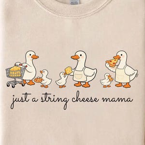Könnte beinhalten: Beigefarbenes Sweatshirt mit einer skurrilen Grafik von Enten, die Lebensmittel wie Pizza und Käse halten. Der Text "just a string cheese mama" steht darunter. Das Sweatshirt hat einen Rundhalsausschnitt und lange Ärmel.