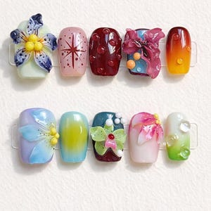 Può includere: Un set di dieci unghie finte con design vari, tra cui decorazioni floreali in 3D, motivi a stella, texture a goccia in rilievo e sfumature di colore nelle tonalità blu, rosa, rosso, giallo, verde e viola.