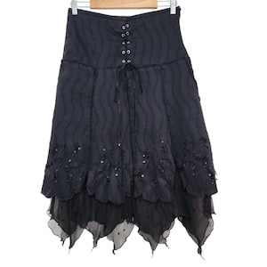 May include: A black midi skirt featuring a high-waisted design with a front lace-up corset detail and metal eyelets. The fabric has a vertical wavy texture, and the hem is layered with a scalloped edge adorned with black sequins and beads.
